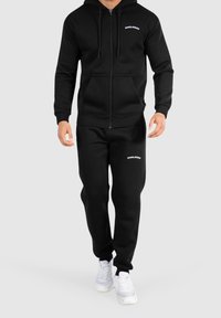 Felpa nera con zip e pantaloni jogger abbinati, realizzati in tessuto morbido, con un logo sul petto e sulla gamba sinistra dei pantaloni, abbinati a sneakers bianche.