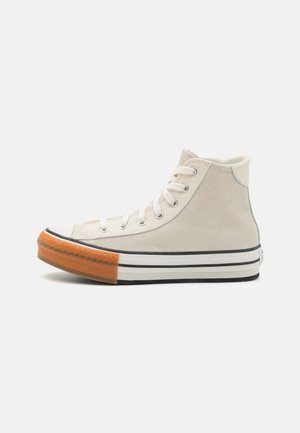 Beżowy sneakers z zamszu za kostkę z białymi sznurowadłami, białą podeszwą w czarne paski i pomarańczowym gumowym noskiem, widziany z boku na białym tle.