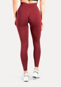 Bordeauxrode legging met hoge taille, een gestructureerd ontwerp en een zijzak, gecombineerd met witte sportschoenen, gezien vanaf de achterkant.