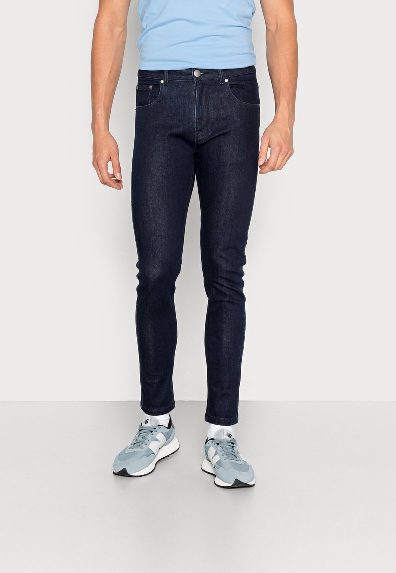 Jeans de denim azul escuro com um corte slim, apresentando cinco bolsos e acessórios metálicos. Usados com uma t-shirt azul clara e sapatilhas cinzentas.