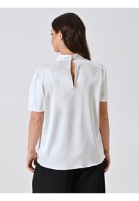 Blusa bianca a maniche corte con colletto alto e apertura a chiave sulla schiena, realizzata in morbido tessuto satinato e con un sottile dettaglio di spalline a palloncino.