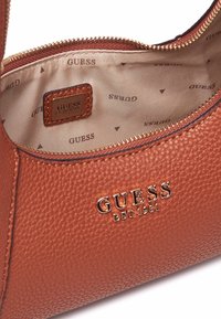 Borsa a mano in pelle marrone con texture, logo dorato, chiusura con zip e fodera beige con motivo ripetuto "GUESS". Etichetta interna visibile.