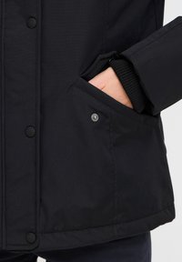 Veste isolante noire avec une texture lisse, dotée d'une fermeture à boutons-pression, de poches latérales et de poignets côtelés.