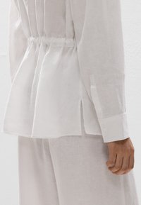Blouse en lin blanc au coupe ample, taille froncée et fentes sur les côtés. Manches longues avec poignets boutonnés, mettant en valeur un tissu doux et texturé.