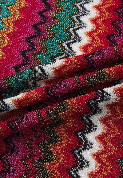 Tejido colorido de fibras entrelazadas que presenta un patrón en zigzag con hilos rojos, negros, blancos, verdes y dorados, mostrando un acabado texturizado.
