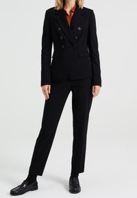 Svart skräddarsydd dubbelknäppt kavaj med fyra runda knappar, i kombination med åtsittande svarta byxor och svarta loafers. Slät tygstruktur.