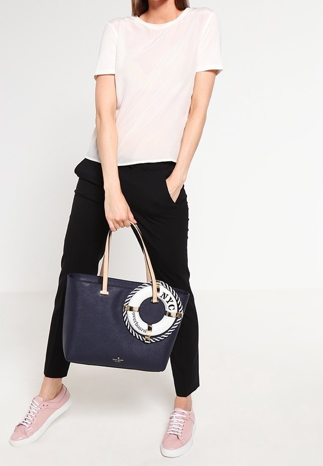 Femme portant un t-shirt blanc, un pantalon noir, des baskets roses, tenant un sac cabas bleu marine avec un porte-clés bouée de sauvetage blanc et bleu marine.