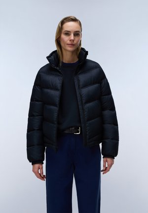 Woman standing wearing a black puffer jacket over a dark sweater and dark blue pants against a plain light gray background.