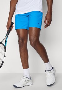 Bright blue swim shorts with an elastic waistband, smooth texture, and a small logo detail. Paired with white sneakers and ankle socks.