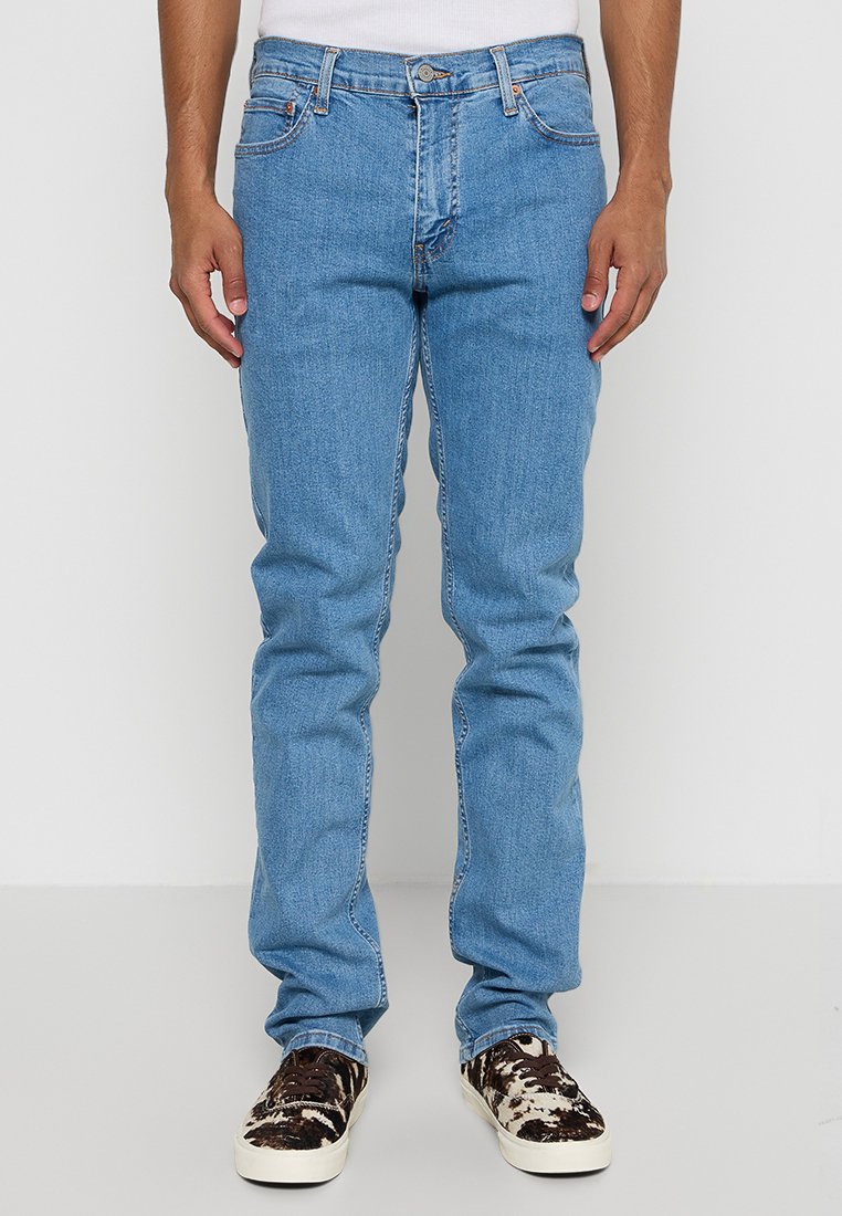 Homme portant un jean droit bleu clair et des baskets blanches avec un motif de vache noir et brun, se tenant devant un fond blanc.