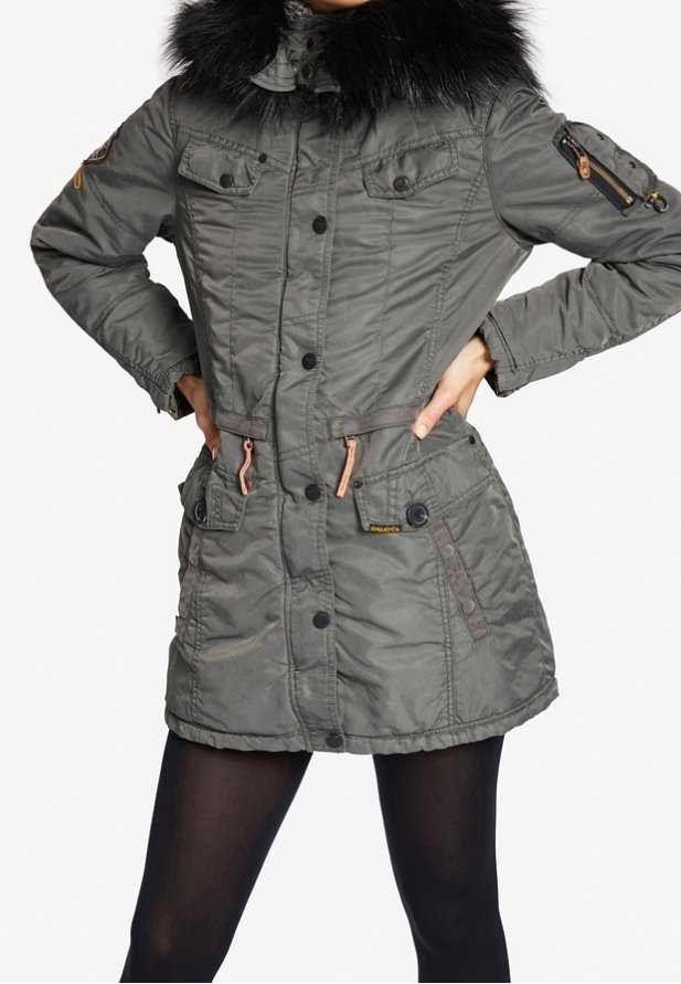 Vrouw in een grijze gewatteerde winterjas met een zwarte bontkraag, knoop- en ritsluiting aan de voorkant, meerdere zakken en zwarte leggings.