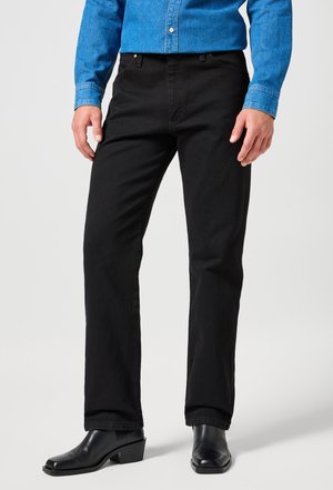 Mann iført svarte bukser med rette ben, svarte skinnstøvler og en blå denim-knappeskjorte, stående foran en ensfarget bakgrunn.