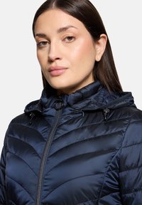 Veste matelassée bleu marine avec motif matelassé, col montant et capuche réglable. Fermeture éclair avec détails en plastique. Texture lisse et brillante.