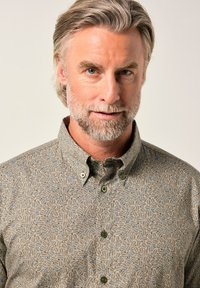 Hombre de mediana edad con cabello y barba gris, vistiendo una camisa abotonada con un patrón de hojas beige y azul, frente a un fondo liso.