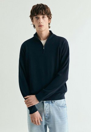 Suéter azul marino con cremallera en tejido suave, con mangas largas y puños acanalados, combinado con vaqueros desgastados de color azul claro. Diseño sencillo y clásico.