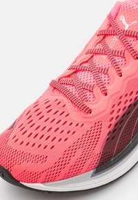 Chaussure de sport rose avec dessus en maille, présentant une finition texturée, des accents noir contrastants et un renfort à l'avant du pied. Les lacets sont intégrés.