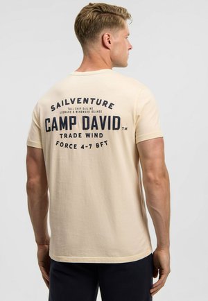 Mann mit kurzen blonden Haaren, von hinten gesehen, trägt ein beiges T-Shirt mit dunkelblauem "CAMP DAVID" segelthemenbezogenem Text auf dem Rücken.