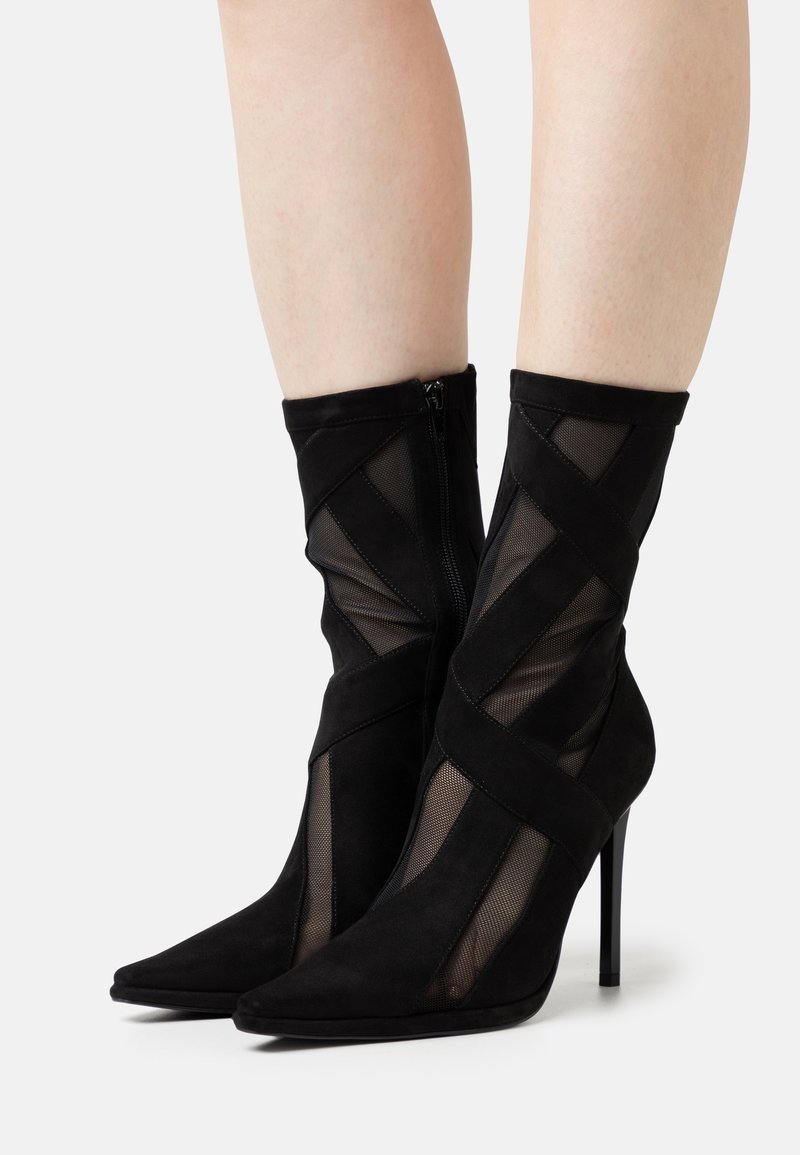 Bottes noires à talons hauts et à cheville, dotées de panneaux en mesh et de brides croisées en daim, portées sur des jambes nues sur un fond blanc.