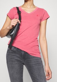 Rosa bomull T-shirt med V-hals, korta ärmar och subtil logotypdetalj, i kombination med mörkgrå jeans och en svart handväska.