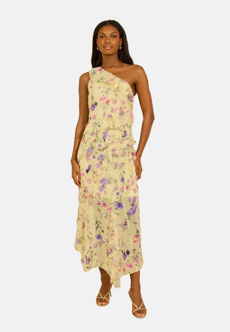 Mujer con un vestido amarillo floral de un solo hombro con volantes y sandalias de tiras, de pie frente a un fondo liso.