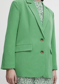Blazer de tela verde con dos botones marrones, diseño de un solo botón, solapas con muesca y un bolsillo delantero. La capa interior presenta una camisa con estampado.