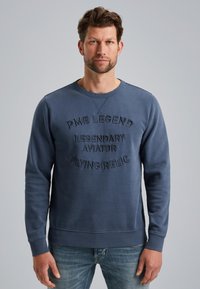 Plavi dukserica s izvezenim tekstom "PME LEGEND LEGENDARY AVIATOR FLYING RELIC", okrugli izrez, te rebrasti rubovi na rukavima i donjem dijelu.