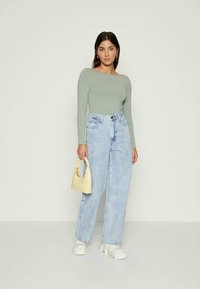 Blusa de manga longa verde-clara combinada com jeans de cintura alta azul-claro. A modelo segura uma pequena mala amarela e usa ténis brancos.