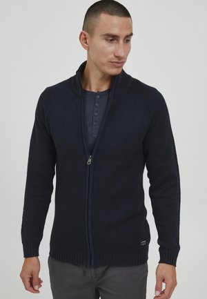 Cárdigan de punto azul marino con cremallera y patrones texturizados, puños y dobladillo acanalados, que presenta un cuello alto y un discreto logo en el lado inferior.