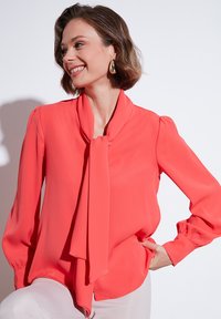 Korallipink blouse, millel on seotud kaeluse detail, pikad varrukad ja sile tekstuur. Kujunduses on kergelt punnitatud õmblused õlgadel.