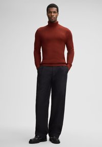 Uomo in piedi con le mani nelle tasche, che indossa un maglione a collo alto testurizzato color ruggine, pantaloni neri a gamba larga e scarpe di pelle nere su uno sfondo grigio.