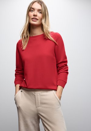 Roter Sweatshirt mit geripptem Halsausschnitt und Bündchen, kombiniert mit hellbeigen Cordhosen, die seitliche Taschen und eine strukturierte Oberfläche aufweisen.
