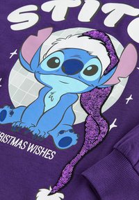 Pull violet avec un extraterrestre bleu en dessin animé portant un bonnet de Père Noël blanc et violet à sequins et le texte « Christmas Wishes » en dessous.
