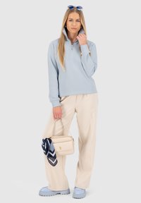 Felpa azzurra con zip, pantaloni ampi color crema, scarpe azzurre. Borsa beige con sciarpa blu e bianca a fantasia attaccata.