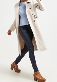 Beige trenchcoat med bälte i midjan, ljusblå topp, mörka skinny jeans och bruna platåskor. Slätt tyg, lång ärm, knapppatchar.