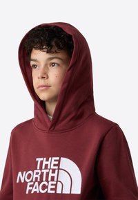 Bordová mikina s kapucňou z mäkkej tkaniny, s veľkým bielym logom na prednej strane. Má podšívanú, nastaviteľnú kapucňu a voľný strih.