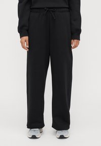Persona che indossa pantaloni neri larghi con coulisse e sneakers grigio chiaro, in piedi contro uno sfondo chiaro e semplice.