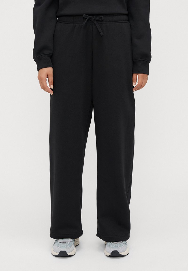Persona che indossa pantaloni neri larghi con coulisse e sneakers grigio chiaro, in piedi contro uno sfondo chiaro e semplice.