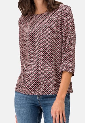 Woman wearing a fitted, patterned long-sleeve top with red and black geometric shapes, paired with blue denim jeans.