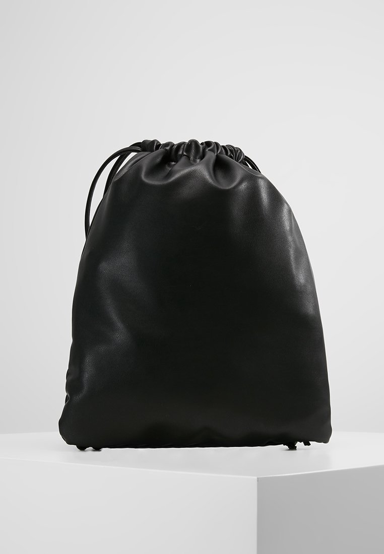 Sacoche à cordon en cuir noir debout sur une surface blanche avec un fond gris clair.