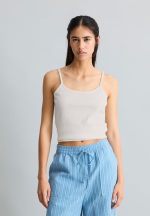 Młoda kobieta z długimi ciemnymi włosami, ubrana w biały prążkowany krótki top i luźne jasnoniebieskie spodnie w paski z troczkiem, stojąca na szarym tle.