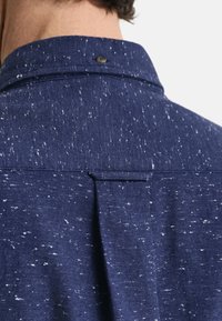 Donkerblauwe shirt van een textuurstof met witte spikkels, een knoopkraag en een rugplooidetail voor extra stijl.