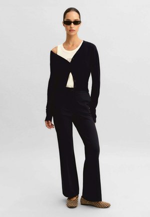 Donna che indossa un cardigan asimmetrico nero sopra una maglietta bianca, pantaloni neri, occhiali da sole scuri e scarpe marroni a pois, in piedi davanti a uno sfondo neutro.