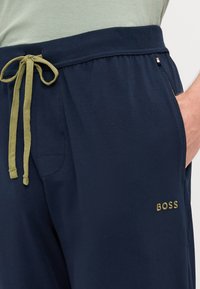 Mornarske trenirke s sproščenim krojem, z olivno zelenim vrvico, stransko žepom in zlatim vezenjem logotipa "BOSS". Gladka tekstura tkanine.