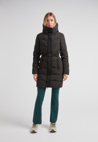Kvinde iført en sort bælte-foret pufferjakke med høj krave, teal bukser og beige sneakers, der står foran en ensfarvet lys baggrund.