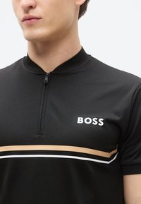 Svart kortermet poloshirt med glidelåslukking i kragen. Har en hvit logo og horisontale striper i beige og hvitt over brystet.