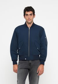 Man wearing a navy blue bomber jacket with zip front, flap pockets, and zippered pocket on the sleeve, paired with faded black jeans.