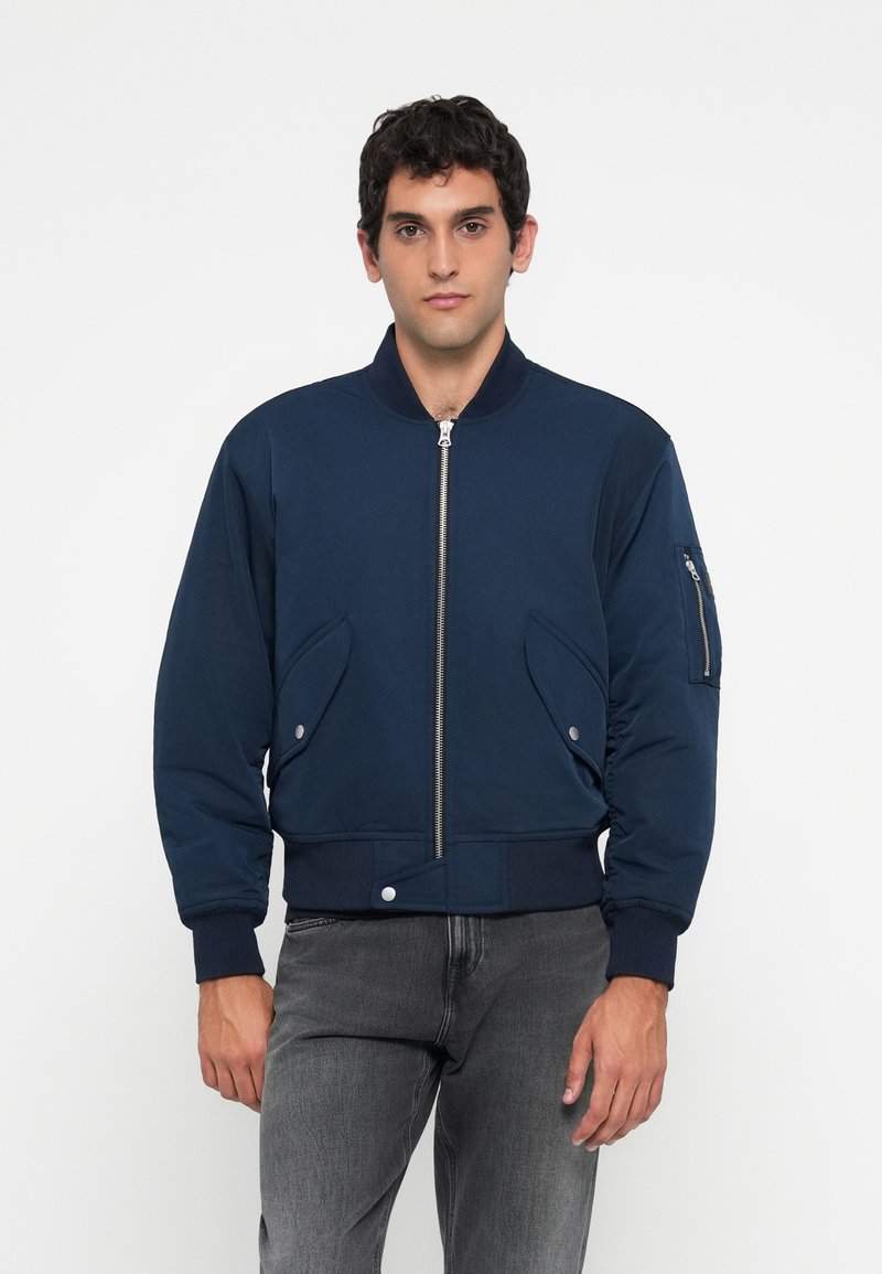 Man wearing a navy blue bomber jacket with zip front, flap pockets, and zippered pocket on the sleeve, paired with faded black jeans.
