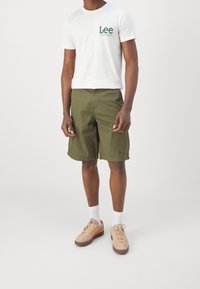 Camiseta blanca con logo verde "Lee", pantalones cortos verde oliva, zapatillas beige con acentos naranjas, calcetines blancos, diseño de cargo simple, corte relajado.