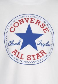 T-shirt blanc arborant un graphique circulaire avec une étoile bleue, du texte rouge "CONVERSE," "ALL STAR," et "Chuck Taylor" dans une police décontractée.