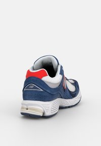 Chaussure de sport New Balance bleue, blanche et rouge avec des panneaux en mesh et en daim, vue de dos et inclinée vers la droite.